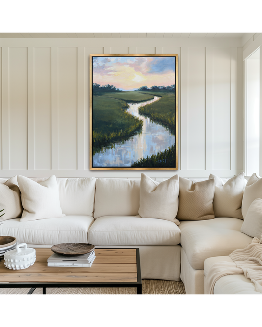 A framed lowcountry marsh landscape painting of a winding tidal river through green fields under a sunset sky, displayed on a cream paneled wall above a white sectional sofa in a modern living room setting.