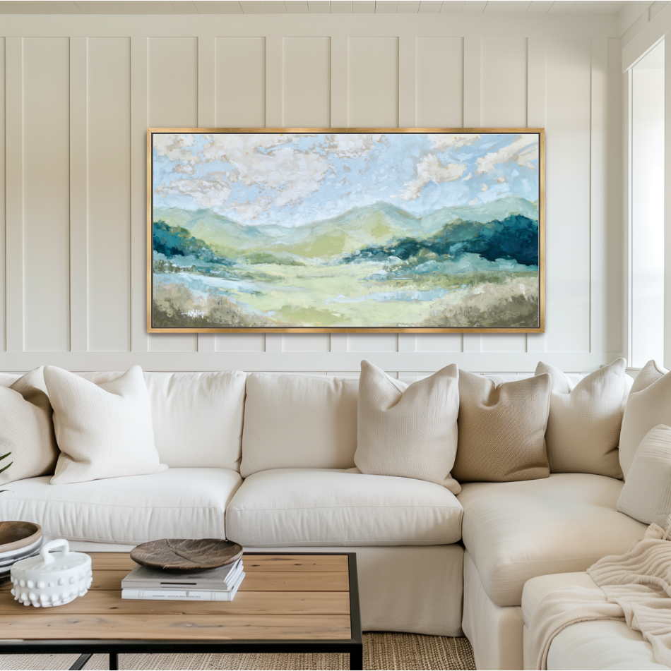 Panoramic Blue Ridge Mountain oil painting in a gold frame mounted above a white sofa.