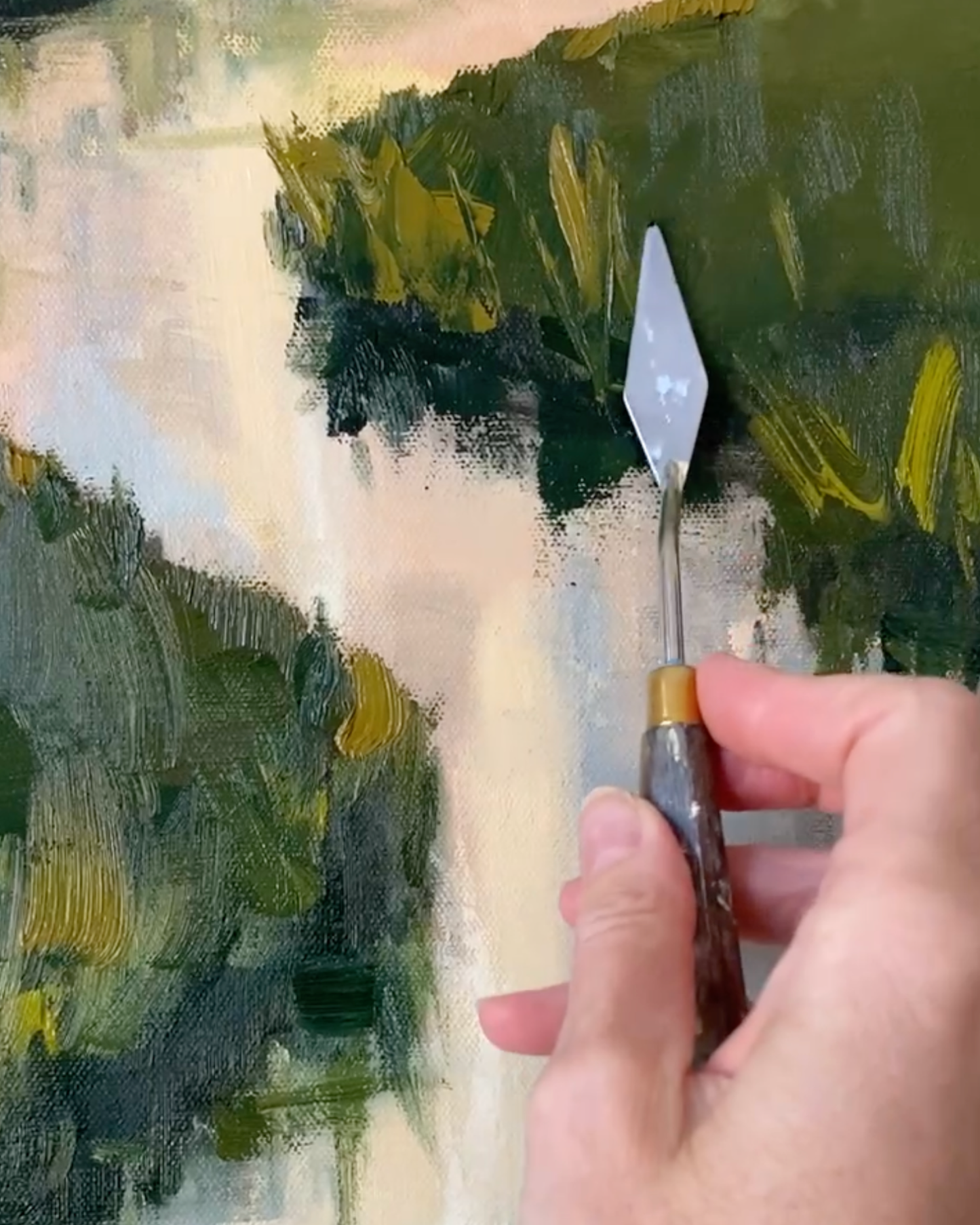 A close-up of a hand holding a palette knife brush with a silver metal blade and brown handle, applying paint to an abstract landscape painting with green, yellow, and cream brushstrokes.