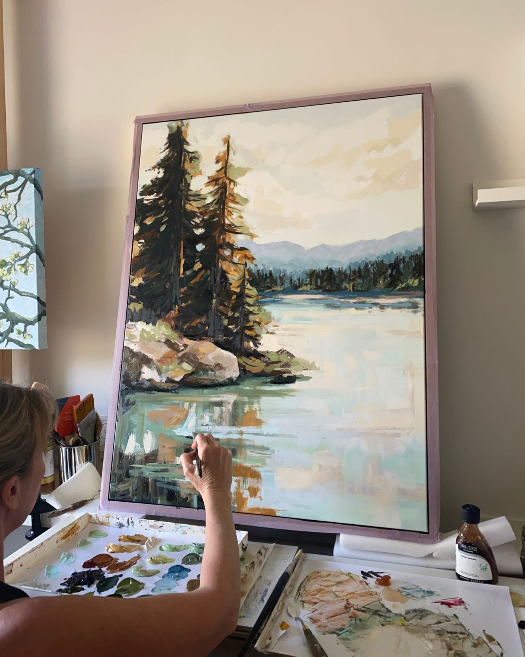 A framed landscape painting of a lake with pine trees and distant mountains, shown in a studio setting with an artist's hand visible in the lower left corner working on the piece in April Moffatt's studio