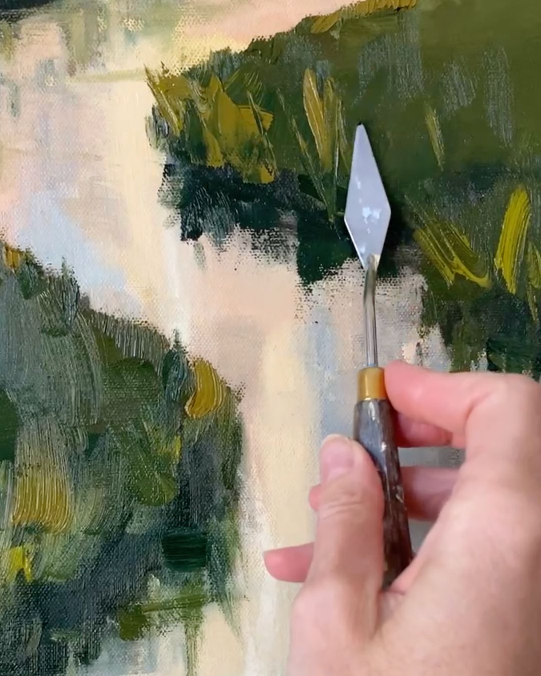 A close-up of a hand holding a palette knife brush with a silver metal blade and brown handle, applying paint to an abstract landscape painting with green, yellow, and cream brushstrokes.