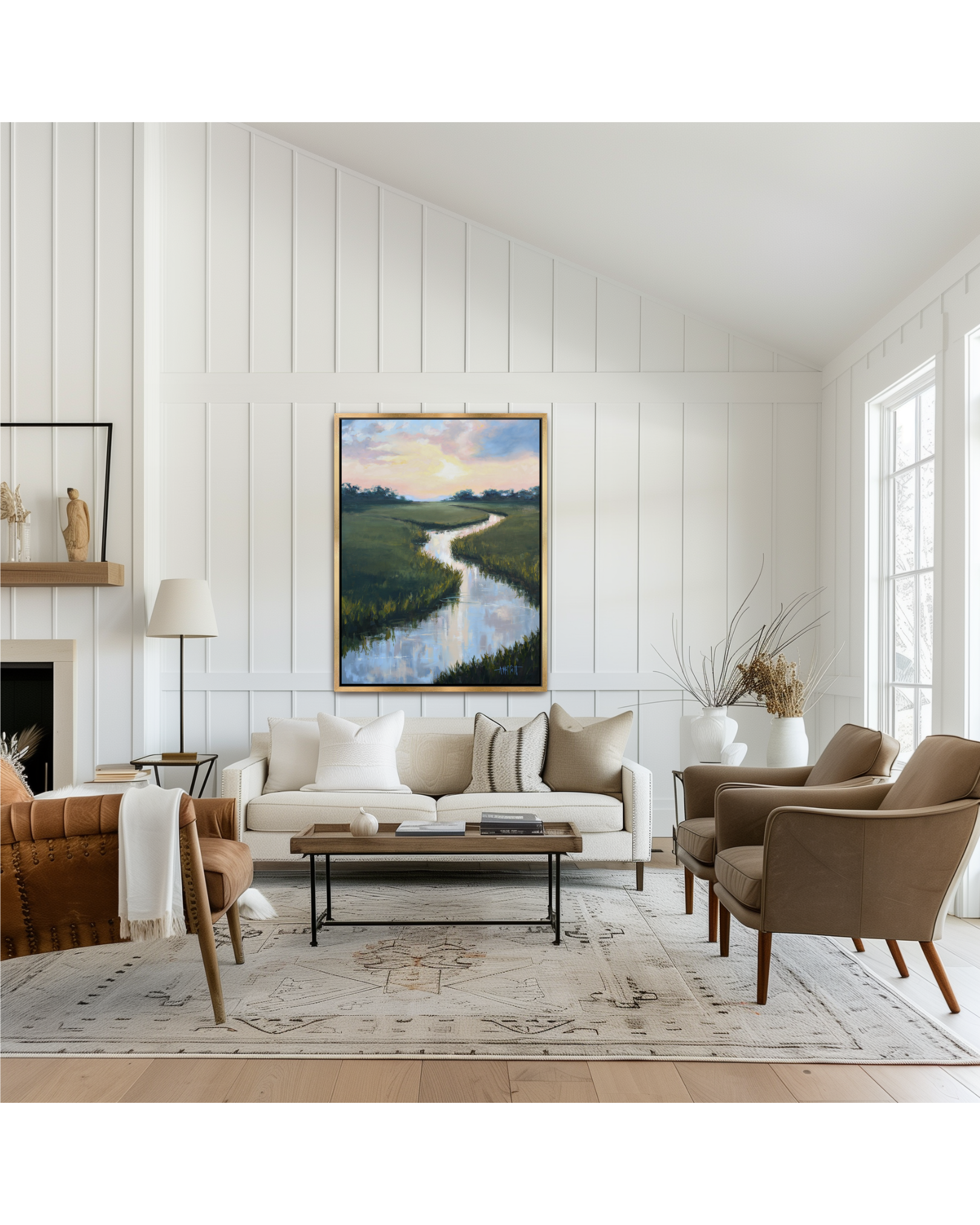 A framed lowcountry marsh landscape painting of a winding river through green fields under a soft sunset sky, displayed on a white paneled wall above a modern coastal living room setup with a sofa, armchairs, and a coffee table.