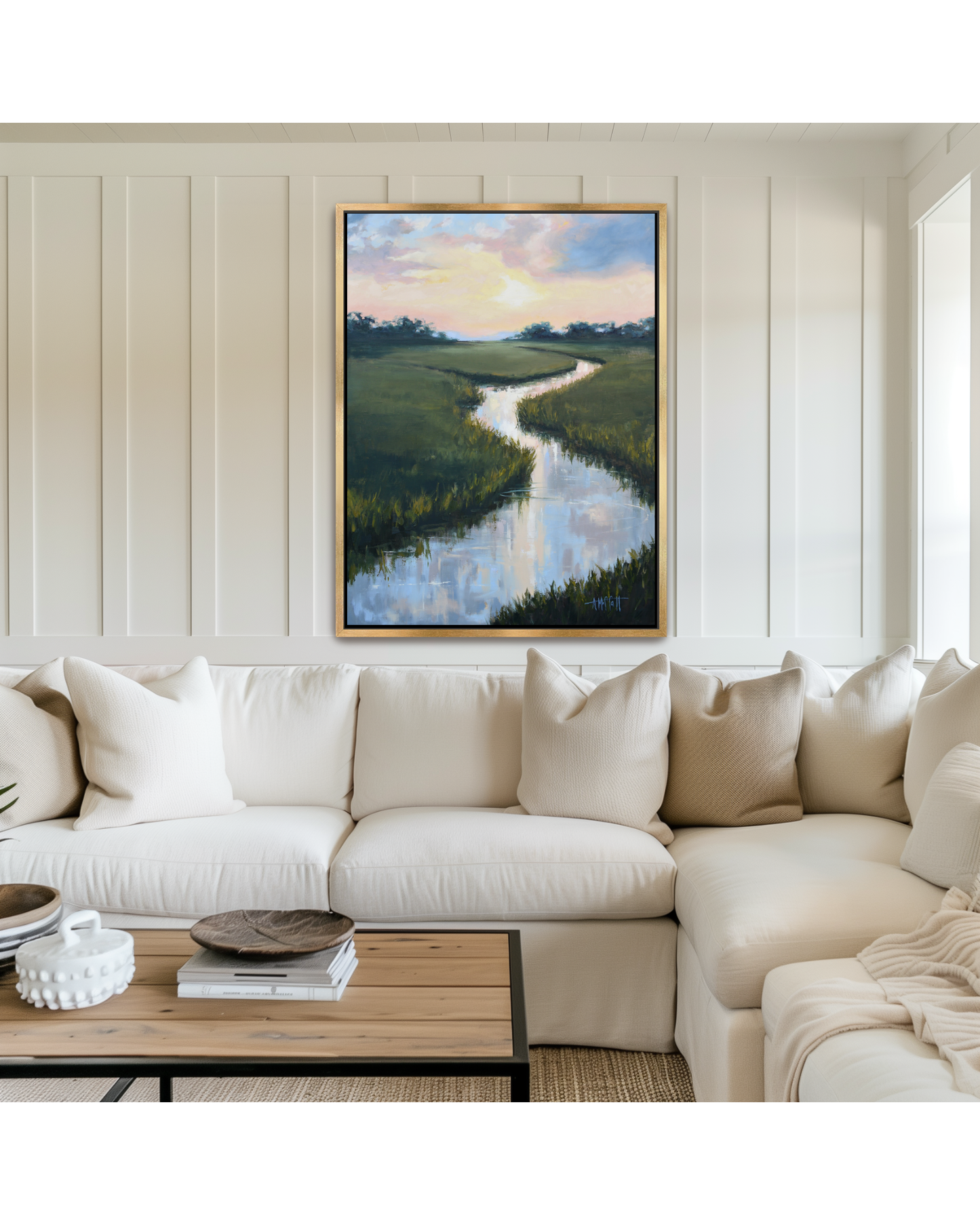 A framed lowcountry marsh landscape painting of a winding tidal river through green fields under a sunset sky, displayed on a cream paneled wall above a white sectional sofa in a modern living room setting.