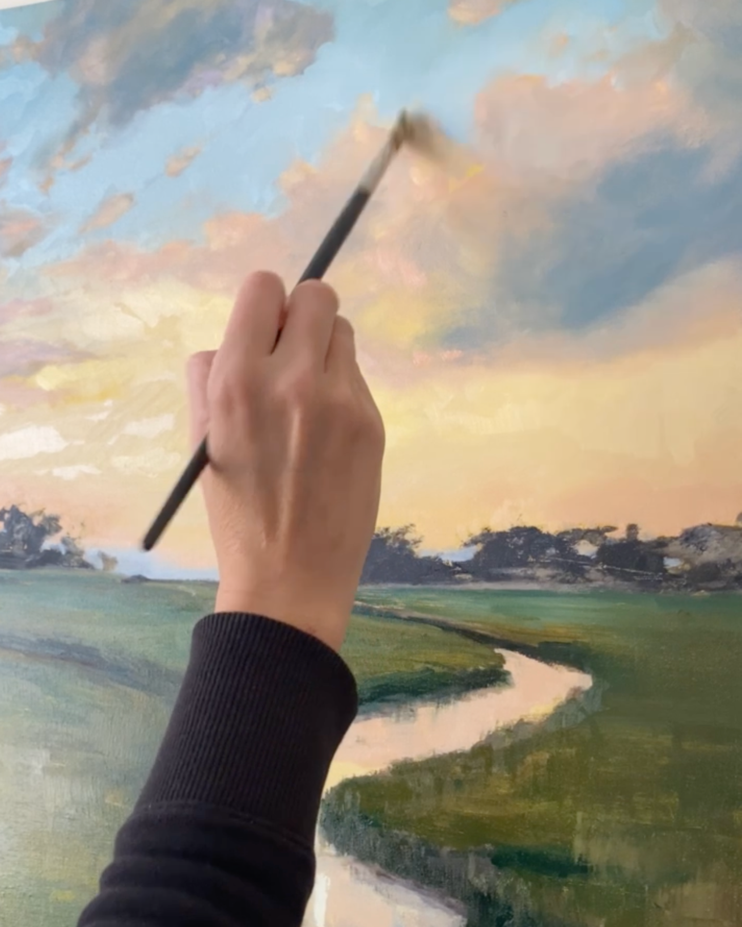 A close-up of April's hand holding a paintbrush and painting a landscape scene with a winding river, green fields, and a cloudy sky.