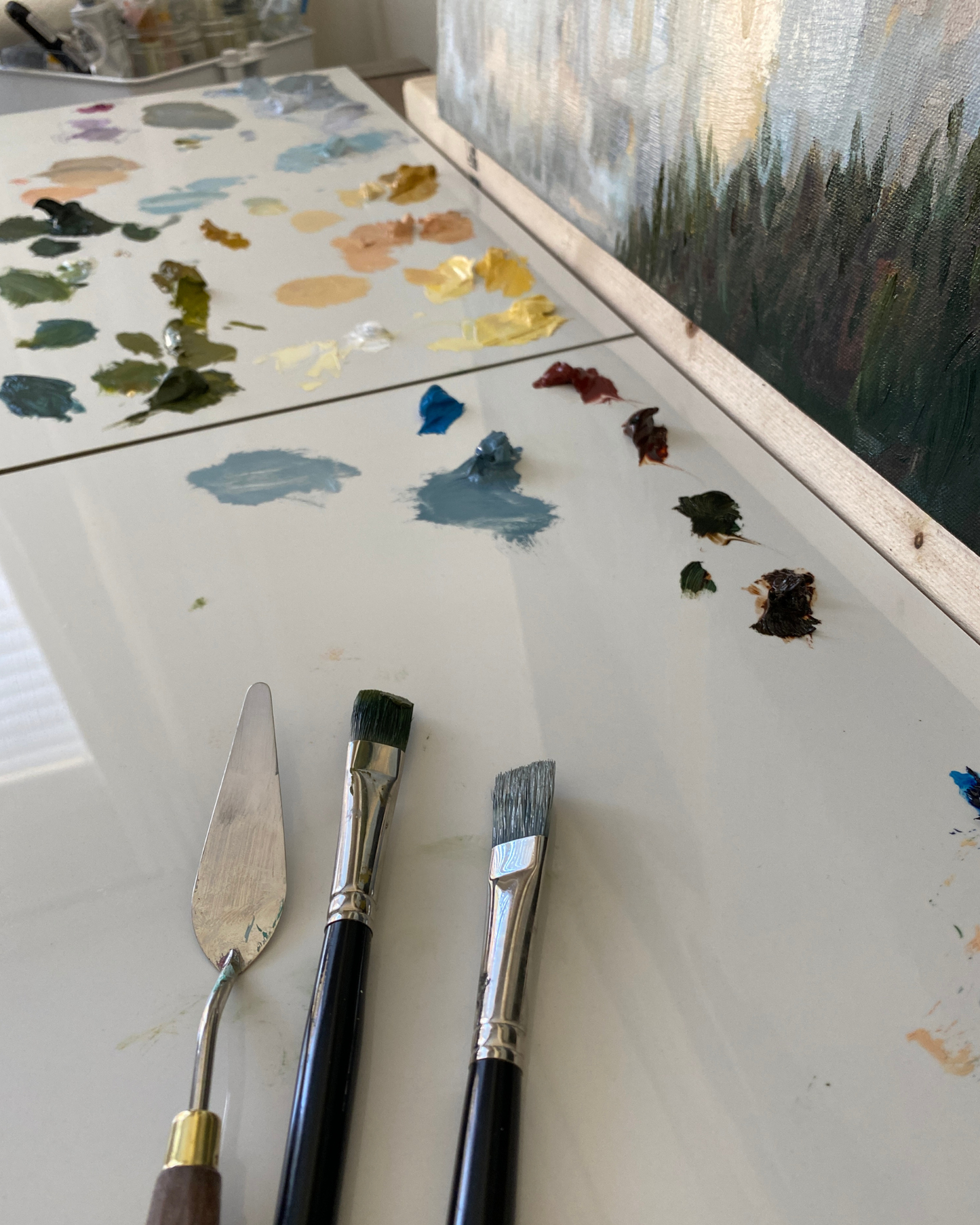 Three painting tools—a metal palette knife, a flat brush, and a round brush—rest on a white artist's palette covered with paint swatches and a canvas showing an impressionistic landscape.
