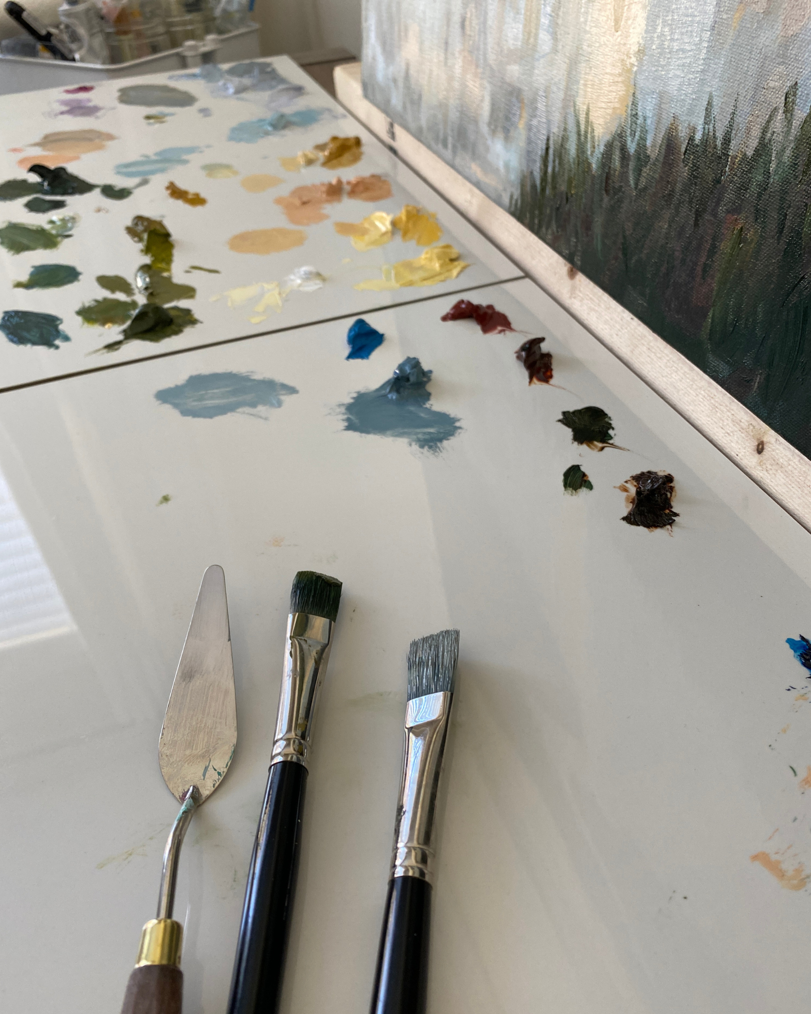 Three painting tools—a metal palette knife, a flat brush, and a round brush—rest on a white artist's palette covered with paint swatches and a canvas showing an impressionistic landscape.